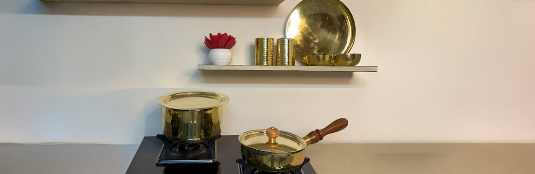 Brass cookware set displayed in a modern kitchen, including brass kadhai, saucepan, glasses, plate, and bowls on shelves and stovetop.